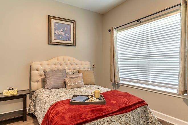 A cozy bedroom with a neatly made bed, featuring decorative pillows and a red throw blanket, complemented by wall art and natural light from the window.