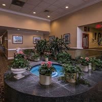 A beautifully decorated lobby features a central fountain surrounded by lush plants and framed artwork.