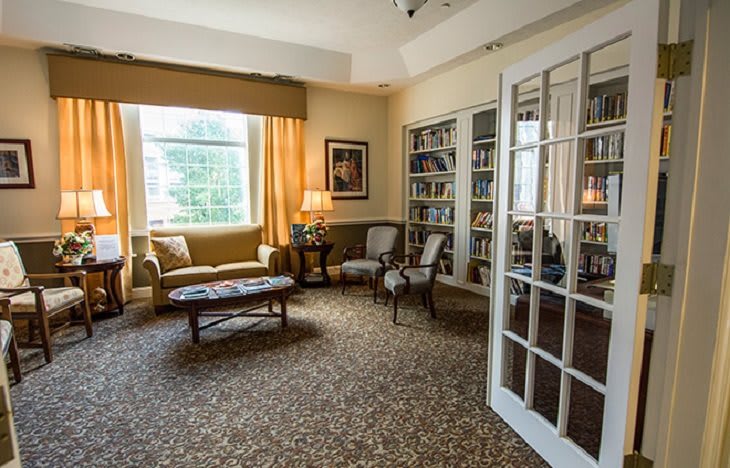 A cozy living room with bookshelves, armchairs, and a large window providing natural light.