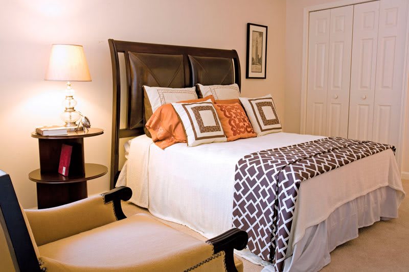 A cozy bedroom with a neatly made bed featuring decorative pillows and a patterned blanket, complemented by warm lighting from a lamp.