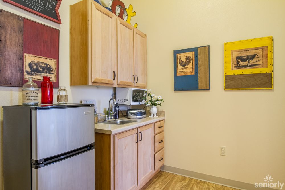 A cozy kitchen corner with wooden cabinets, a small countertop, and various appliances including a mini fridge and microwave. Colorful artwork adorns the walls.