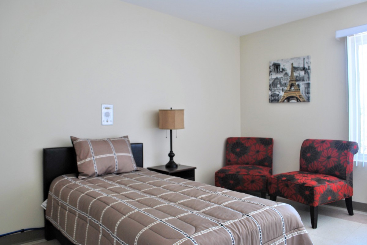 A cozy bedroom with a single bed, a lamp on the nightstand, and two patterned chairs.