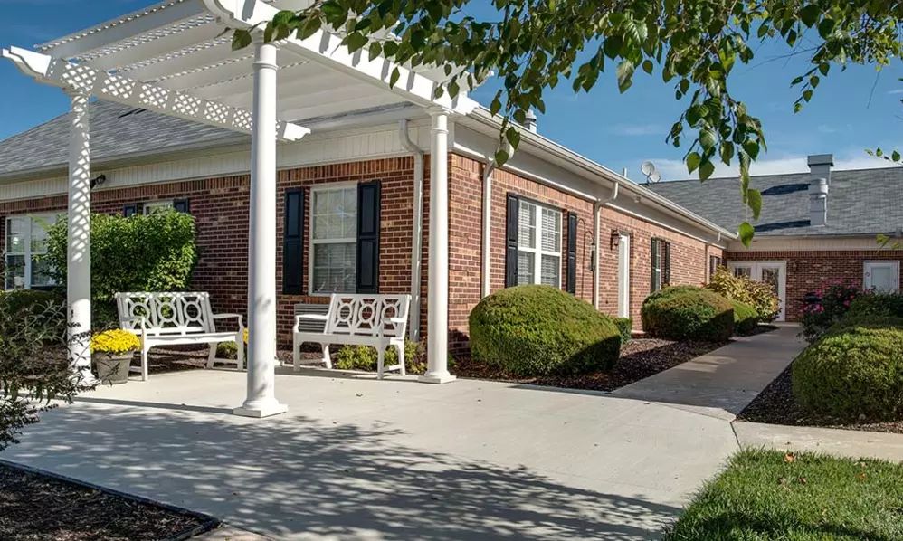 A brick building with a covered porch and benches, surrounded by well-maintained shrubbery.