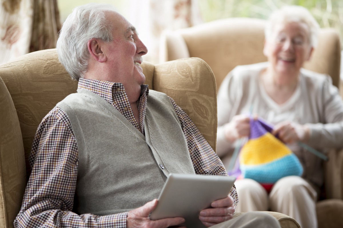 An elderly couple enjoys a moment together, with the man using a tablet while smiling at his partner.