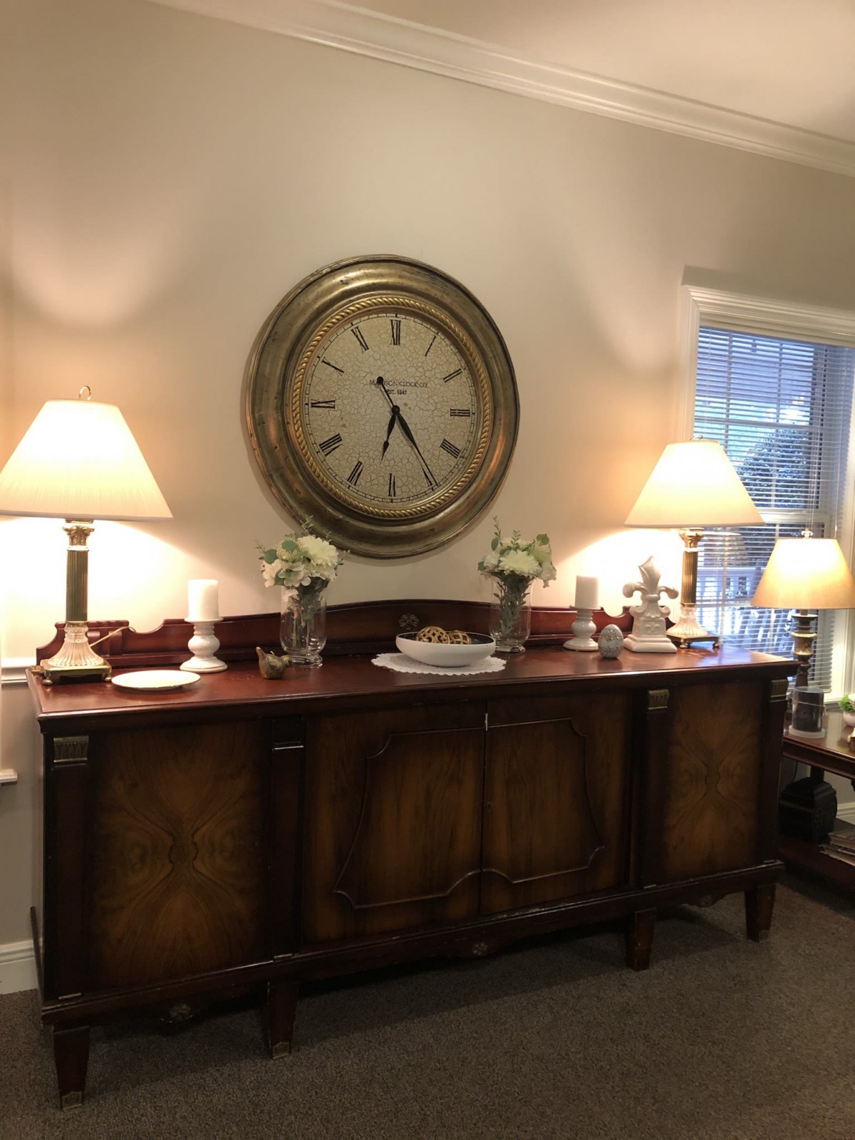 A cozy living room setup featuring a sideboard with decorative lamps and a large wall clock.