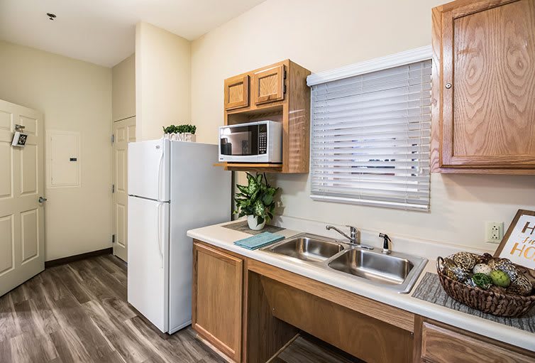 A compact kitchen with essential appliances, featuring a refrigerator, microwave, dual sink, and wooden cabinets.