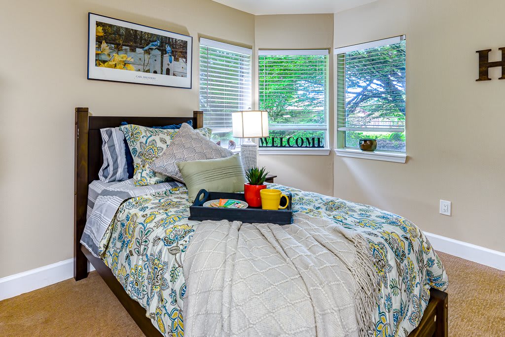 A cozy bedroom with a neatly made bed, featuring decorative pillows and a tray with items on it. The room is well-lit by natural light coming through multiple windows.