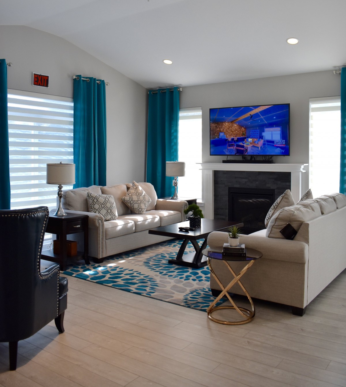 A modern living room with neutral-colored sofas, a wall-mounted television above a fireplace, and vibrant teal curtains.