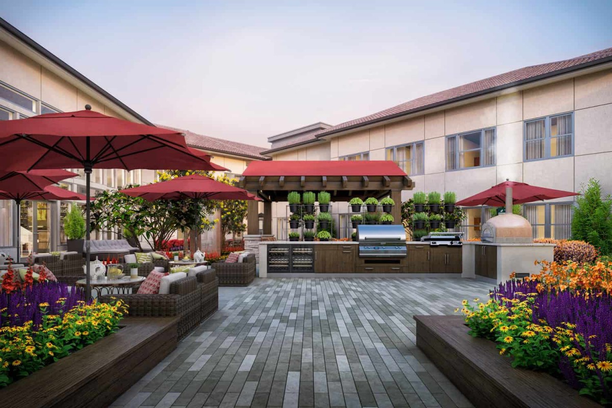 A spacious outdoor patio with dining tables, red umbrellas, and a grill area surrounded by plants.
