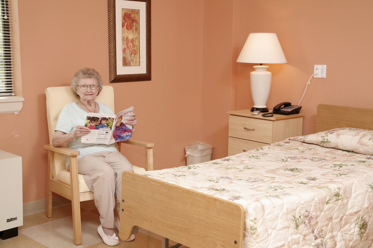 An elderly woman sits in a comfortable chair reading in a cozy bedroom.