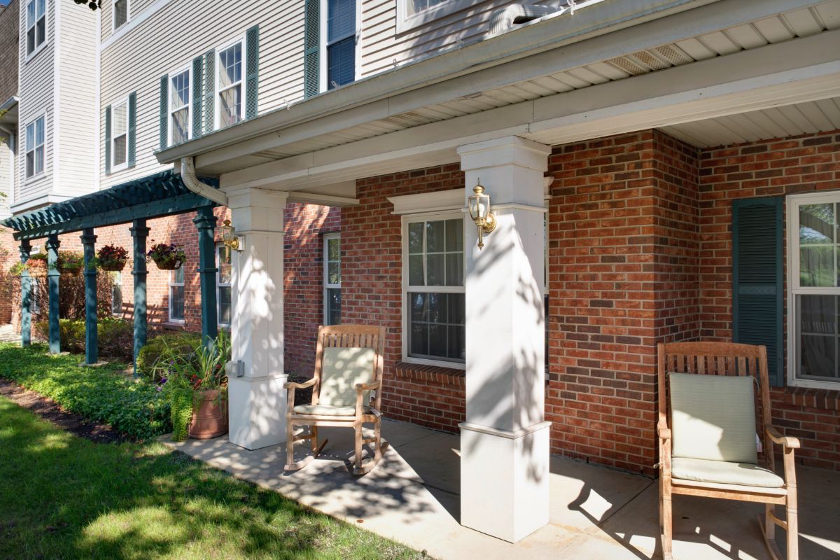 A charming house porch with brick walls and decorative columns, featuring two chairs basking in the sunlight.