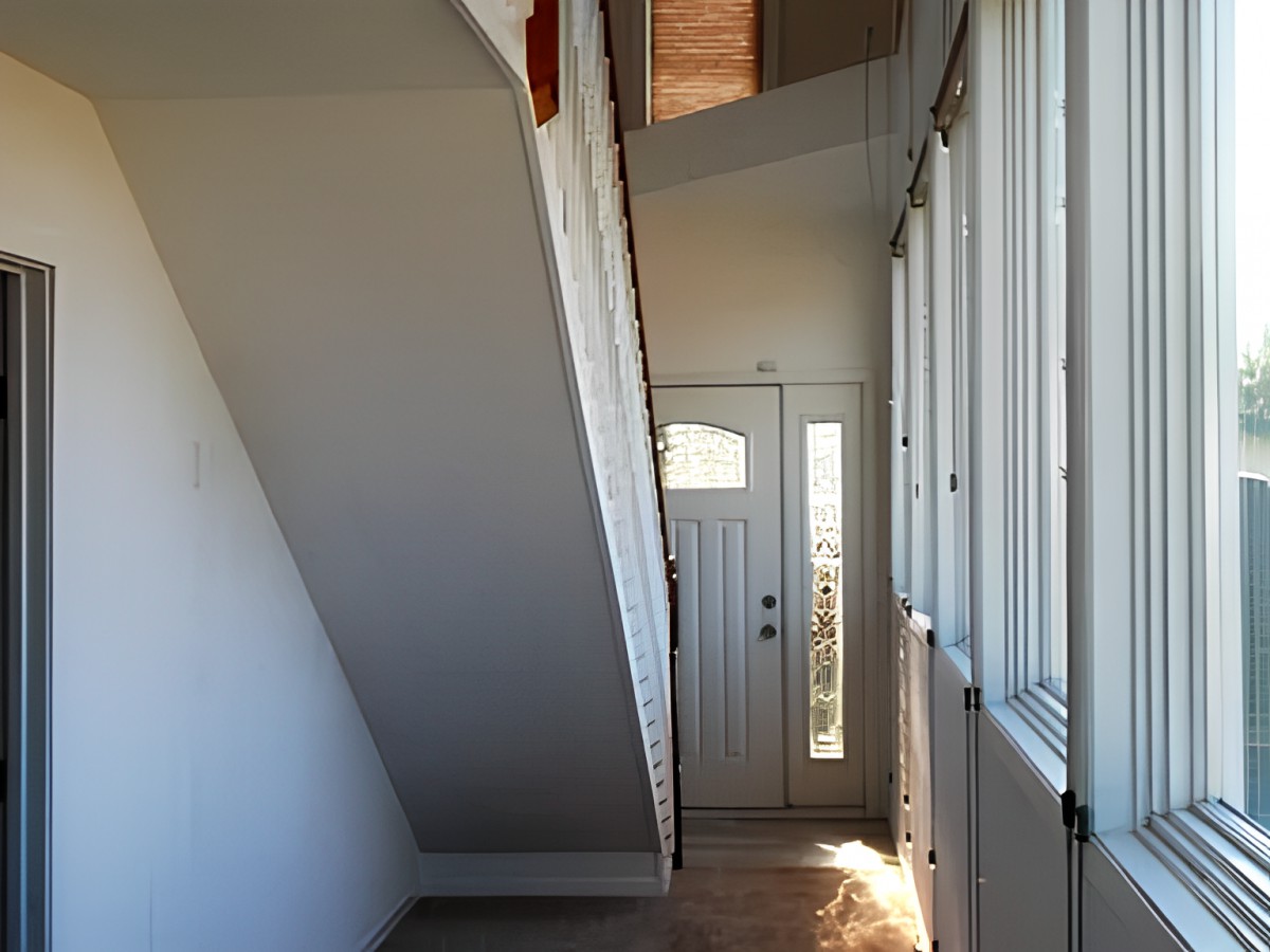 A narrow hallway with a staircase above and a front door at the end, illuminated by natural light through side windows.