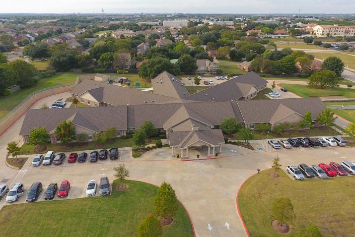 Aerial view of a large building complex surrounded by a parking lot and green landscape in a suburban area.