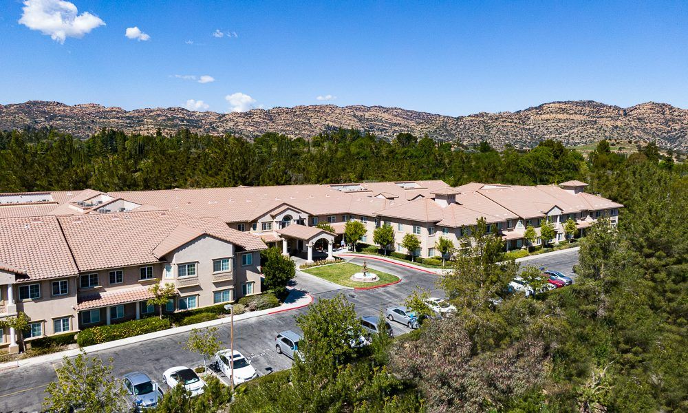 Aerial view of a large building with a parking lot surrounded by trees and mountains under a clear blue sky.