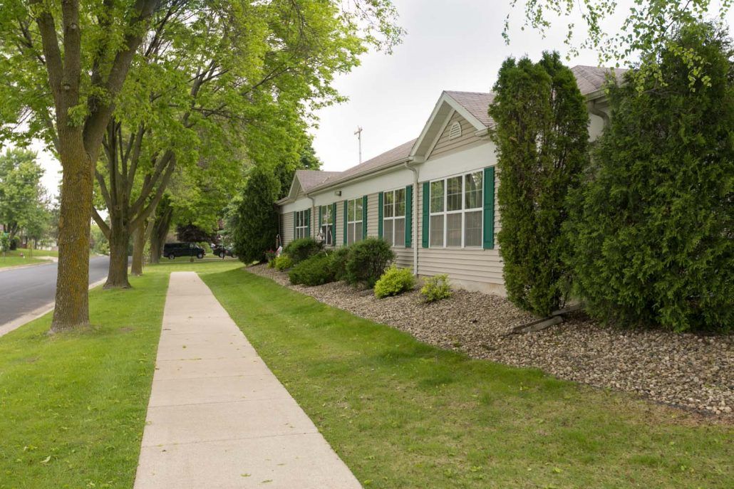 A sidewalk lined with trees and bushes leads past a residential house.