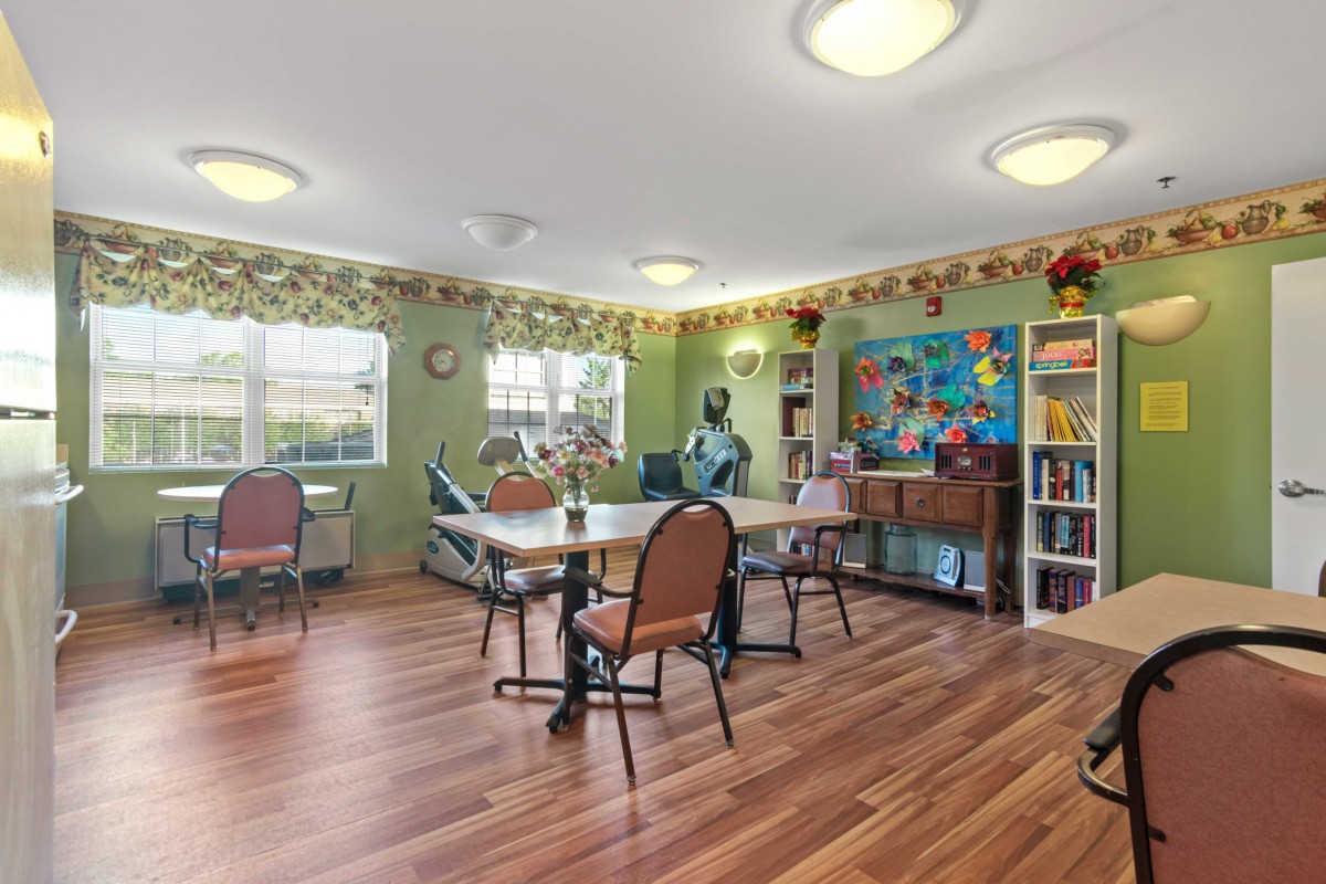 A well-lit community room with tables and chairs, decorated with colorful art and a bookshelf.