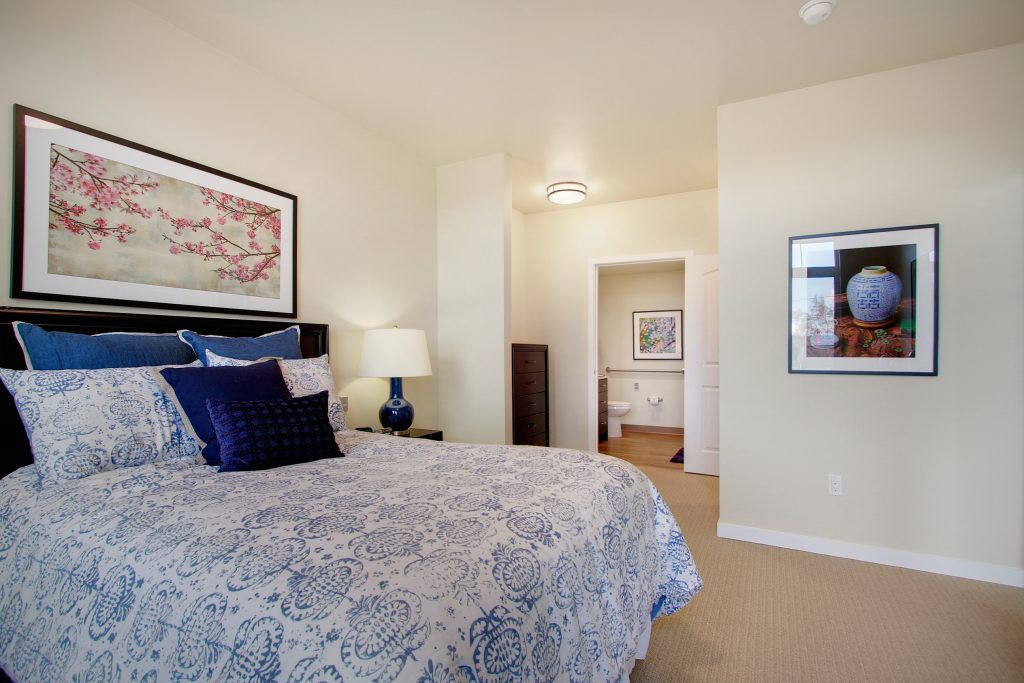A cozy bedroom featuring a bed with patterned bedding, artwork on the walls, and a lamp on the nightstand.