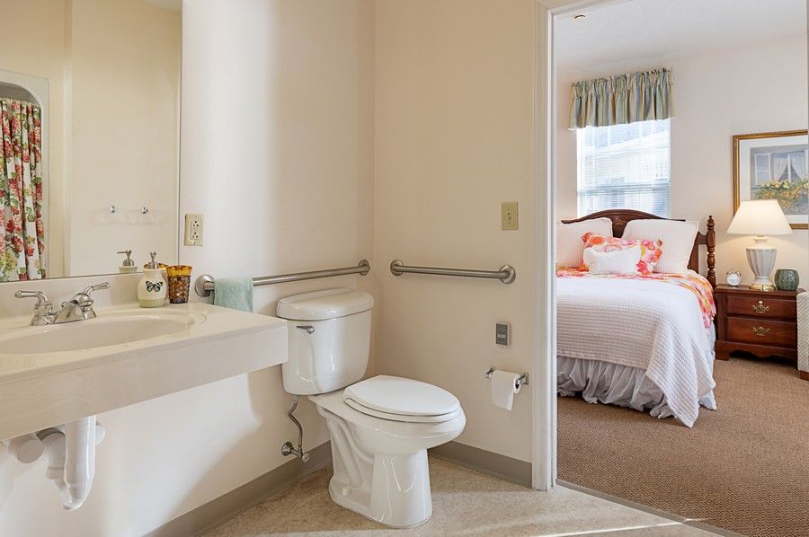 A bathroom with a toilet, sink, and grab bars, adjacent to a bedroom.