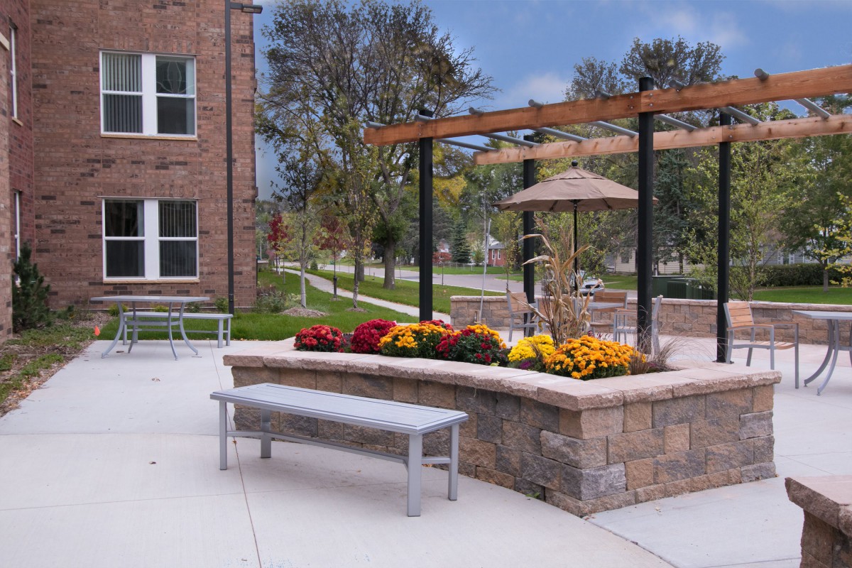 A serene outdoor patio area with a bench, flower bed, and pergola near a brick building.