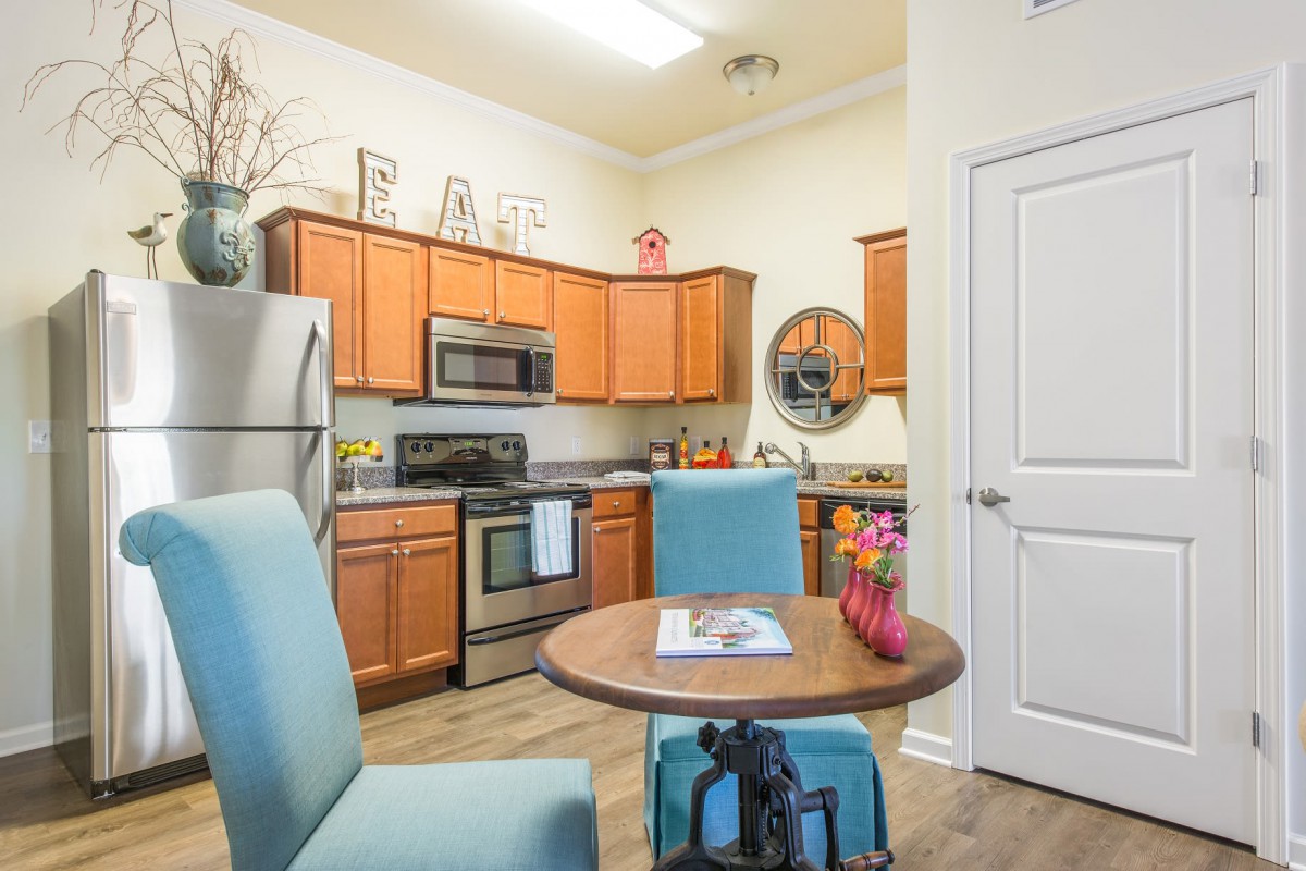 A cozy kitchen with wooden cabinets, stainless steel appliances, and a small dining table with blue chairs.