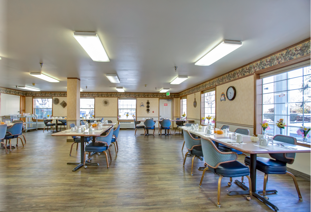 A spacious dining area with arranged tables and chairs, featuring natural lighting and a wooden floor.