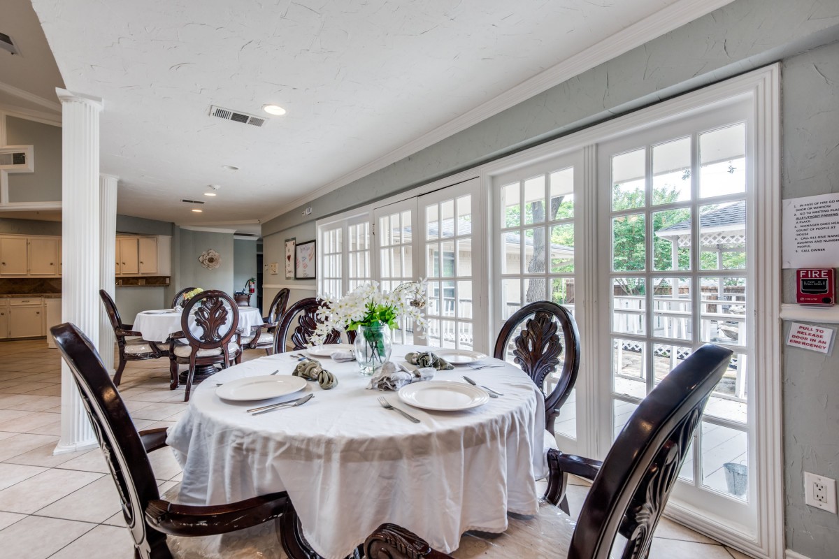 An elegantly set dining room with a round table, surrounded by chairs and illuminated by natural light from large French doors.