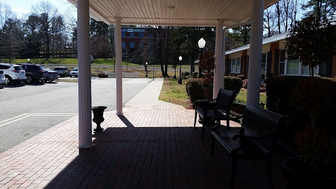 A shaded porch area with benches overlooks a sunlit pathway lined with lamp posts.