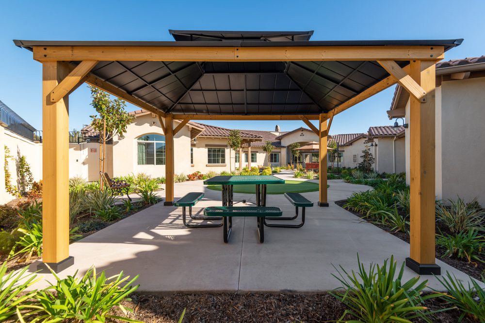 A wooden gazebo with a green picnic table underneath, surrounded by a landscaped garden on a sunny day.