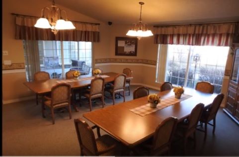 A cozy dining room with large tables and chairs, illuminated by overhead lighting and natural light from the windows.