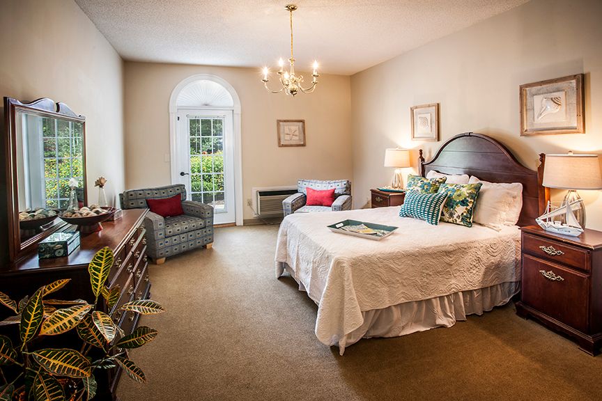 A cozy and elegantly furnished bedroom featuring a bed with decorative pillows, a chandelier, and armchairs by the window.