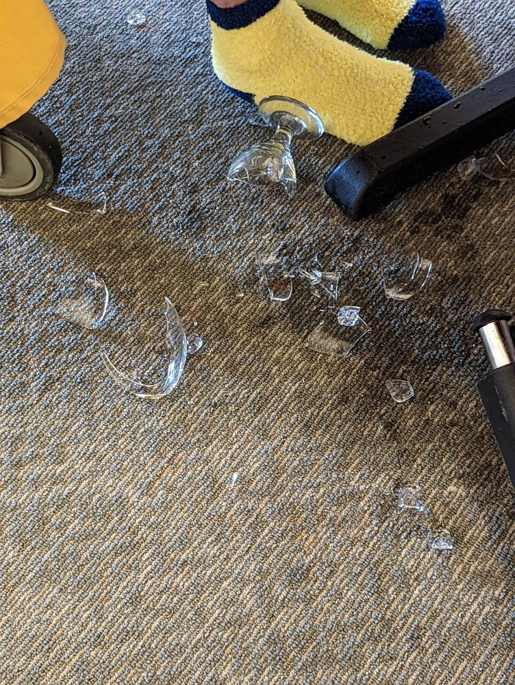 Broken glass and spilled contents on an office carpet near a chair.