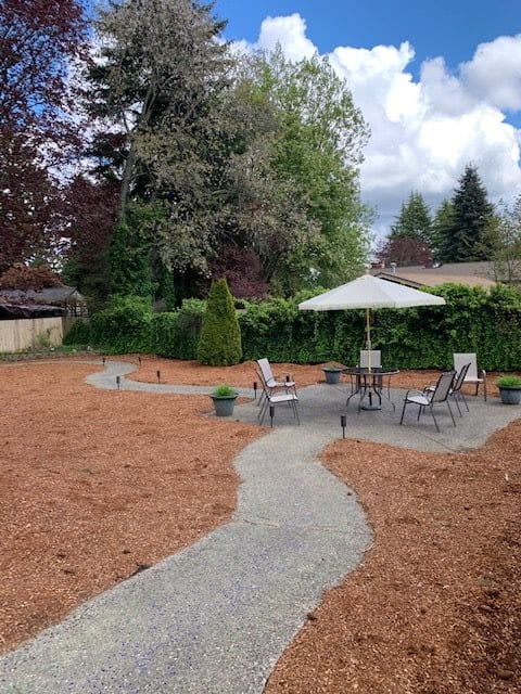 A cozy backyard patio with seating and an umbrella, surrounded by trees and mulch.