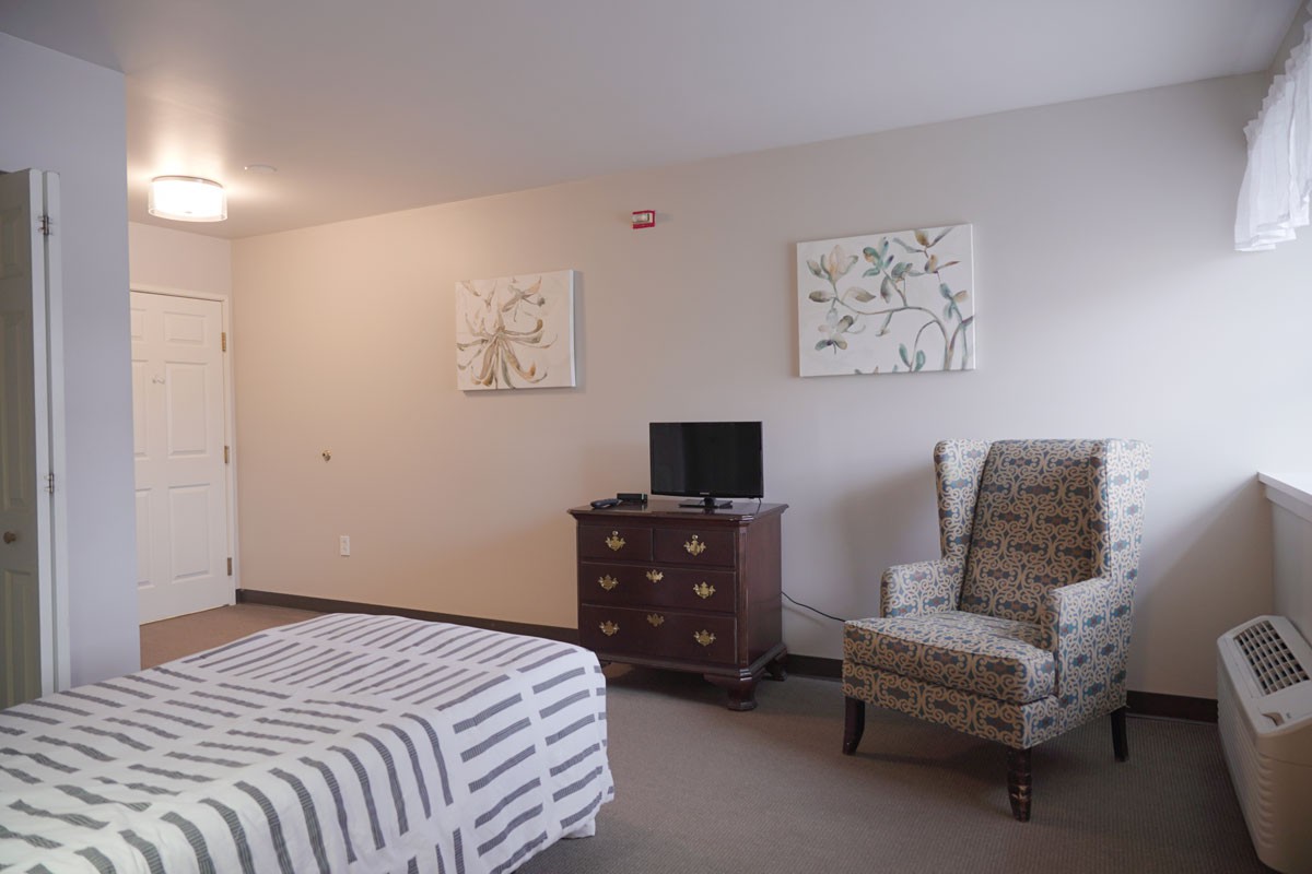 A cozy bedroom featuring a bed, patterned chair, dresser with a television, and decorative paintings on the wall.
