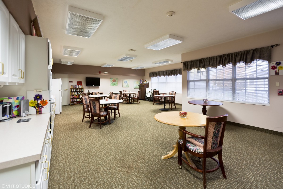 A spacious room with several tables and chairs, carpeted flooring, and large windows. The room is well-lit with ceiling lights, and there are bookshelves along the walls.