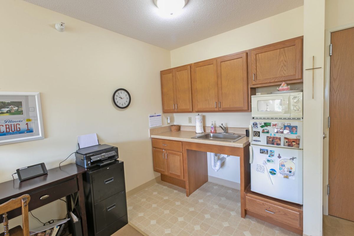 A small kitchen with wooden cabinets, a fridge, microwave, and a clock on the wall.