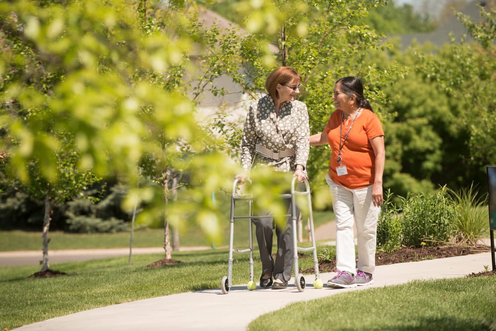 A caregiver assists an elderly person with a walker outdoors on a sunny day.