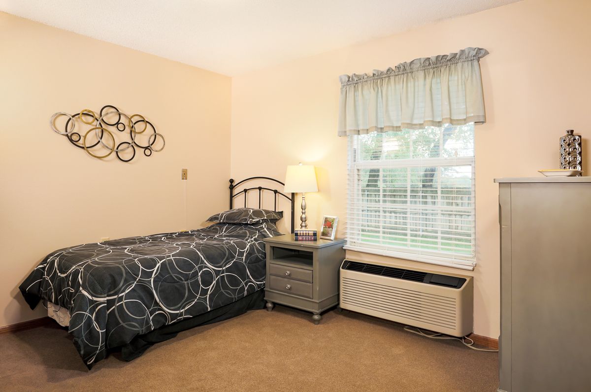 A cozy bedroom with a single bed, nightstand and lamp beside a window adorned with curtains.