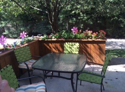 A cozy outdoor patio with a glass table and chairs, surrounded by lush plants and vibrant flowers.