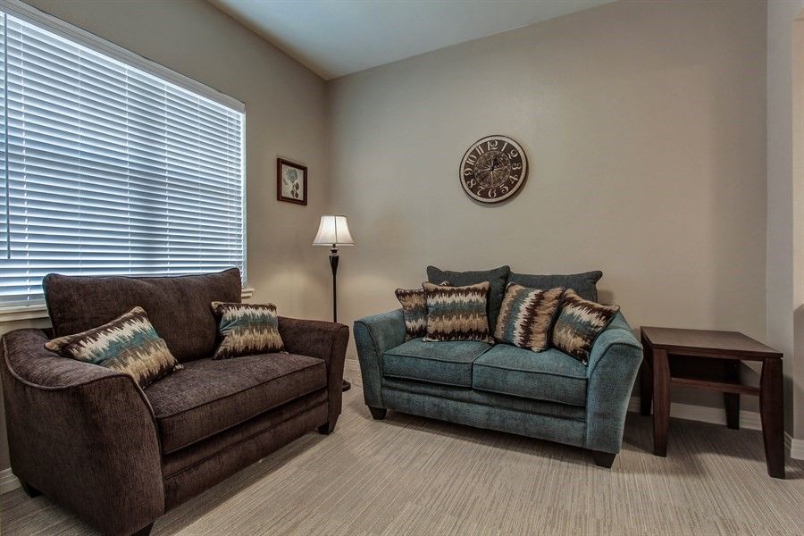 A cozy living room with a brown and blue sofa set, decorative pillows, a floor lamp, and window blinds.