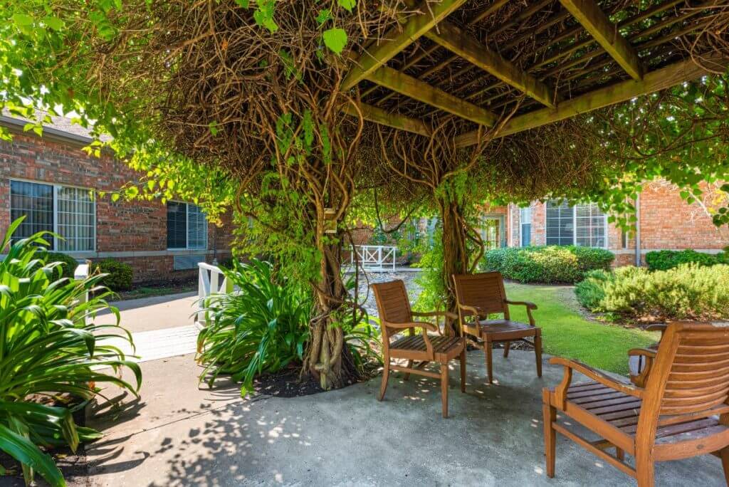 A cozy outdoor gazebo surrounded by lush vines and greenery, featuring wooden chairs for relaxation.
