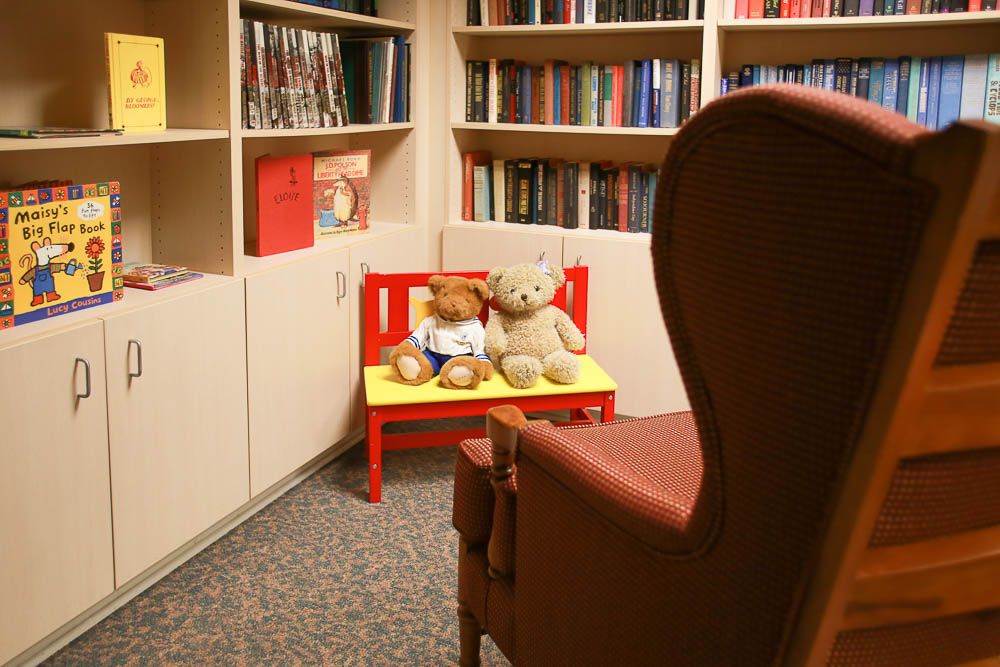 A cozy library corner with teddy bears on a small red bench surrounded by shelves of books.