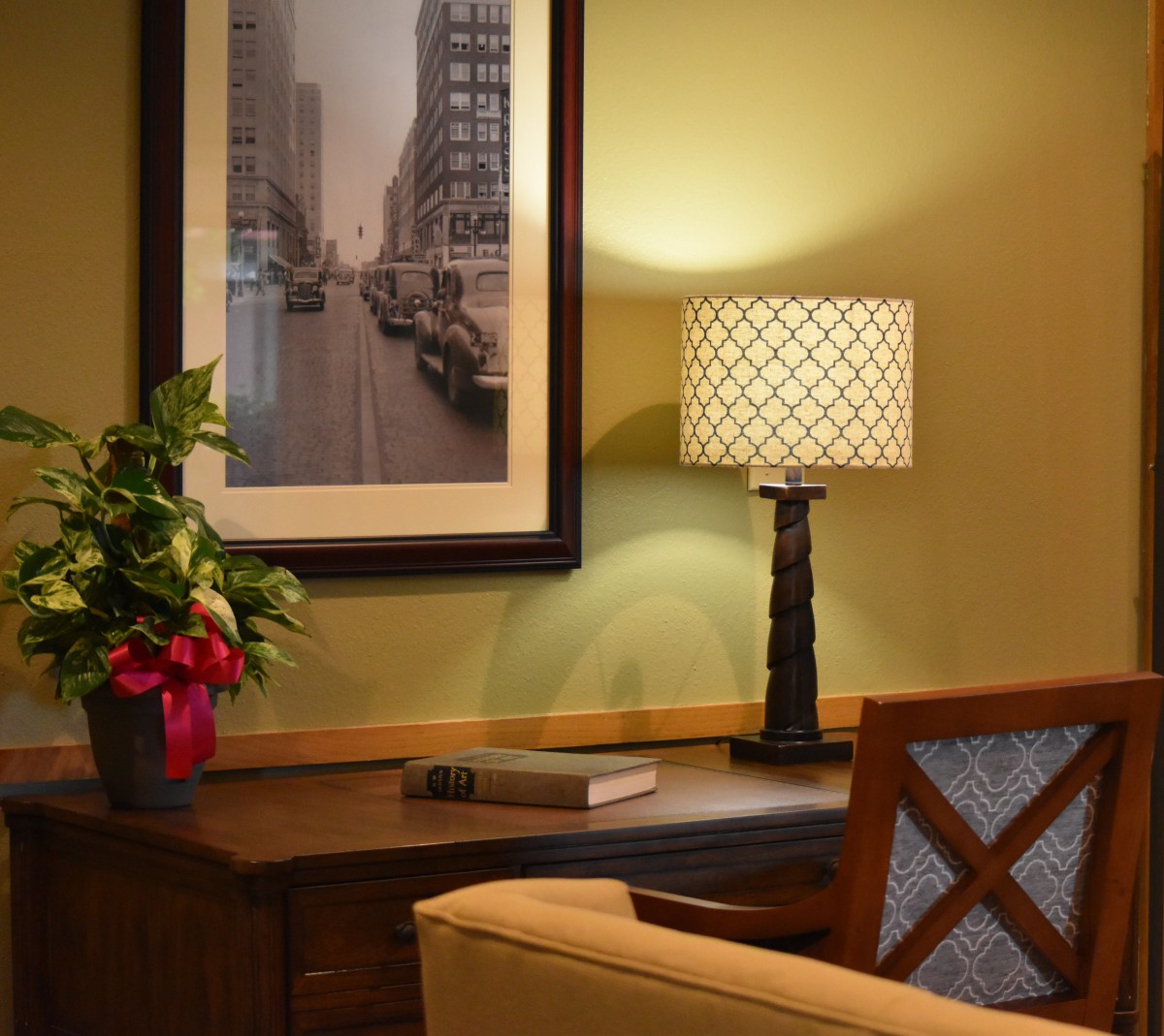 A cozy home office with a wooden desk, decorative lamp, framed cityscape photo, and potted plant.