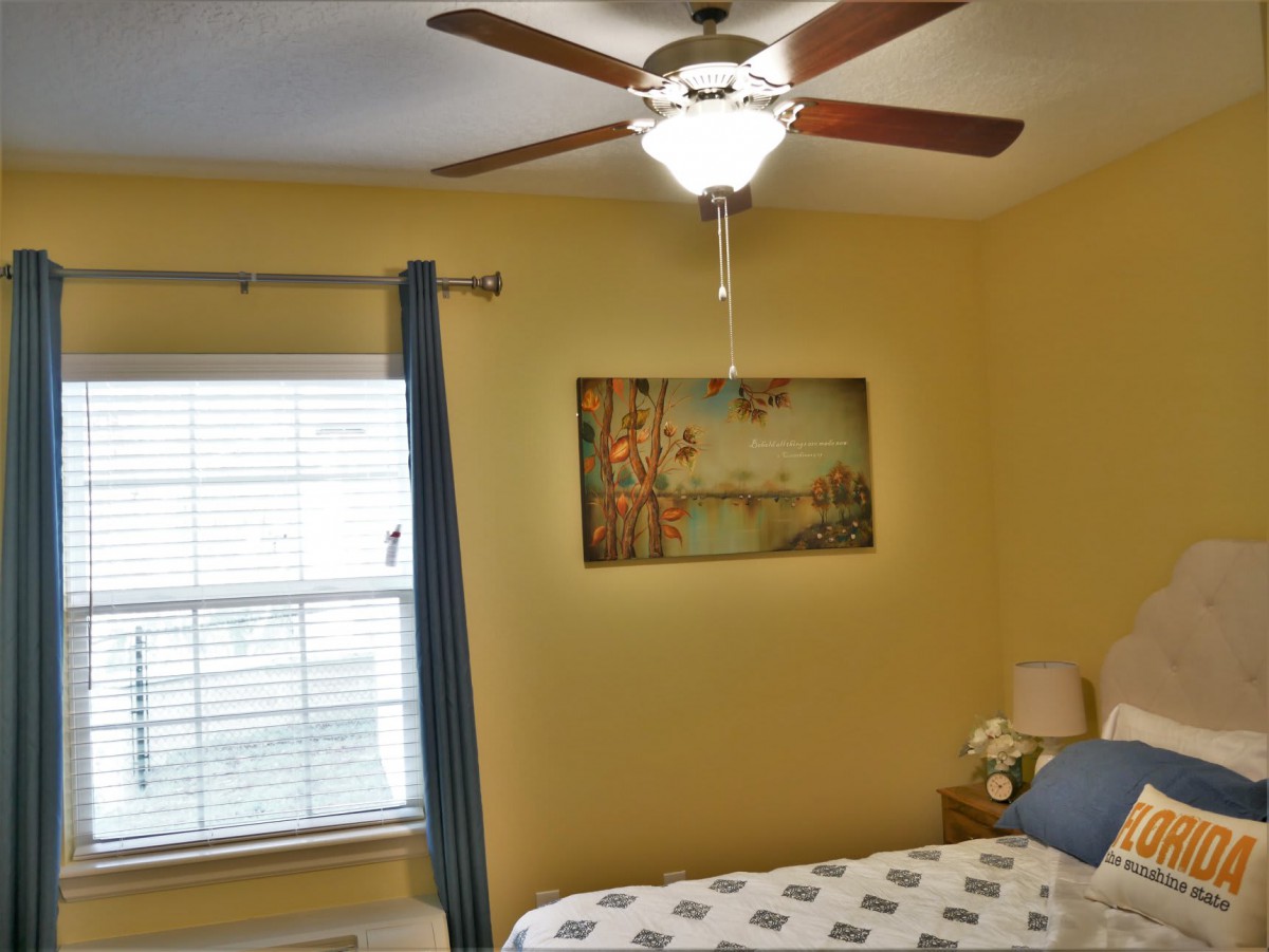 A cozy bedroom with a ceiling fan, window, curtains, and a painting above the bed.