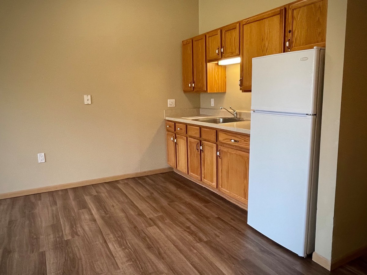 An empty apartment kitchen featuring wooden cabinets, a white refrigerator, and laminate flooring.