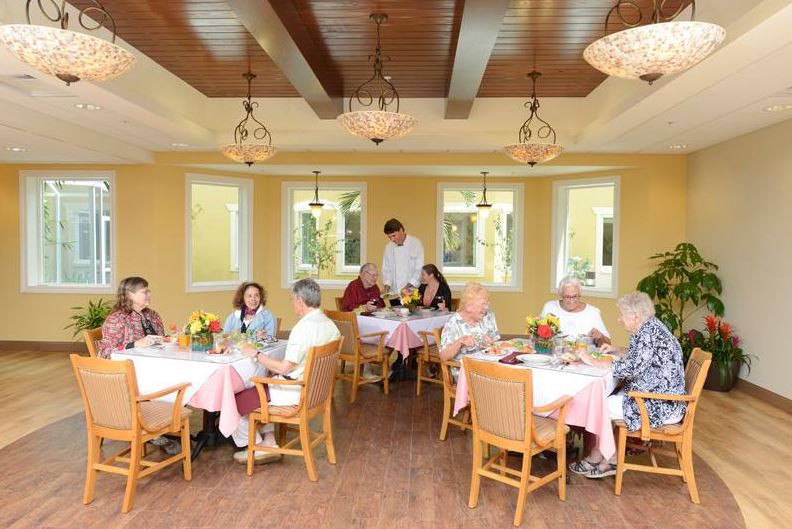 A cozy dining room where seniors are enjoying a meal, served by a waiter.