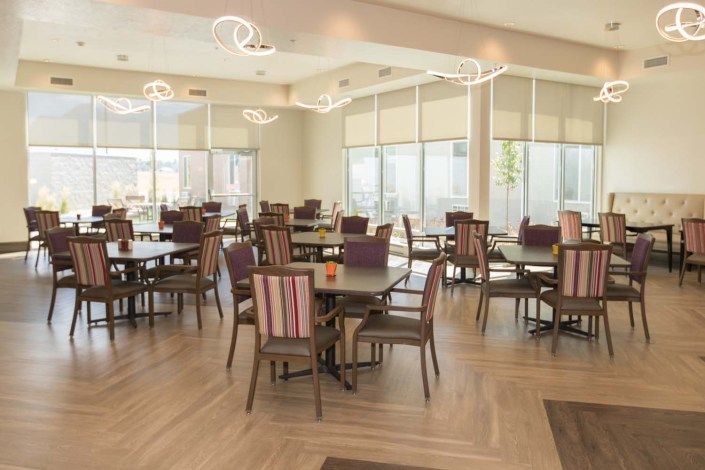 A modern dining area with tables and chairs, illuminated by natural sunlight through large windows.