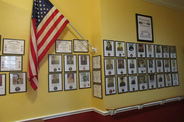 A wall display featuring an American flag, certificates, and framed photos arranged on a yellow background.