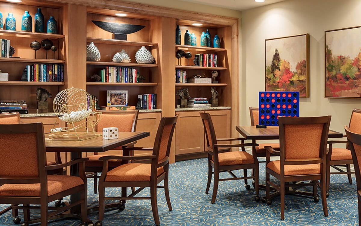 A cozy library setting with bookshelves, tables, and chairs, decorated with various items and board games.