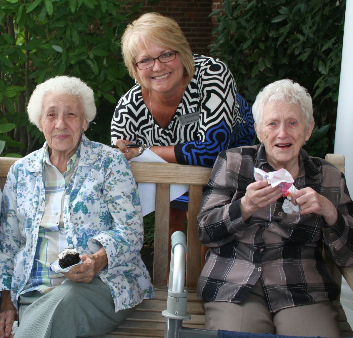 Three women enjoying an outdoor gathering, sitting on a bench and smiling.