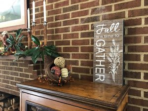 A cozy fall-themed mantel display with candles, a basket of pumpkins, and a wooden 'Fall Gather' sign against a brick wall.